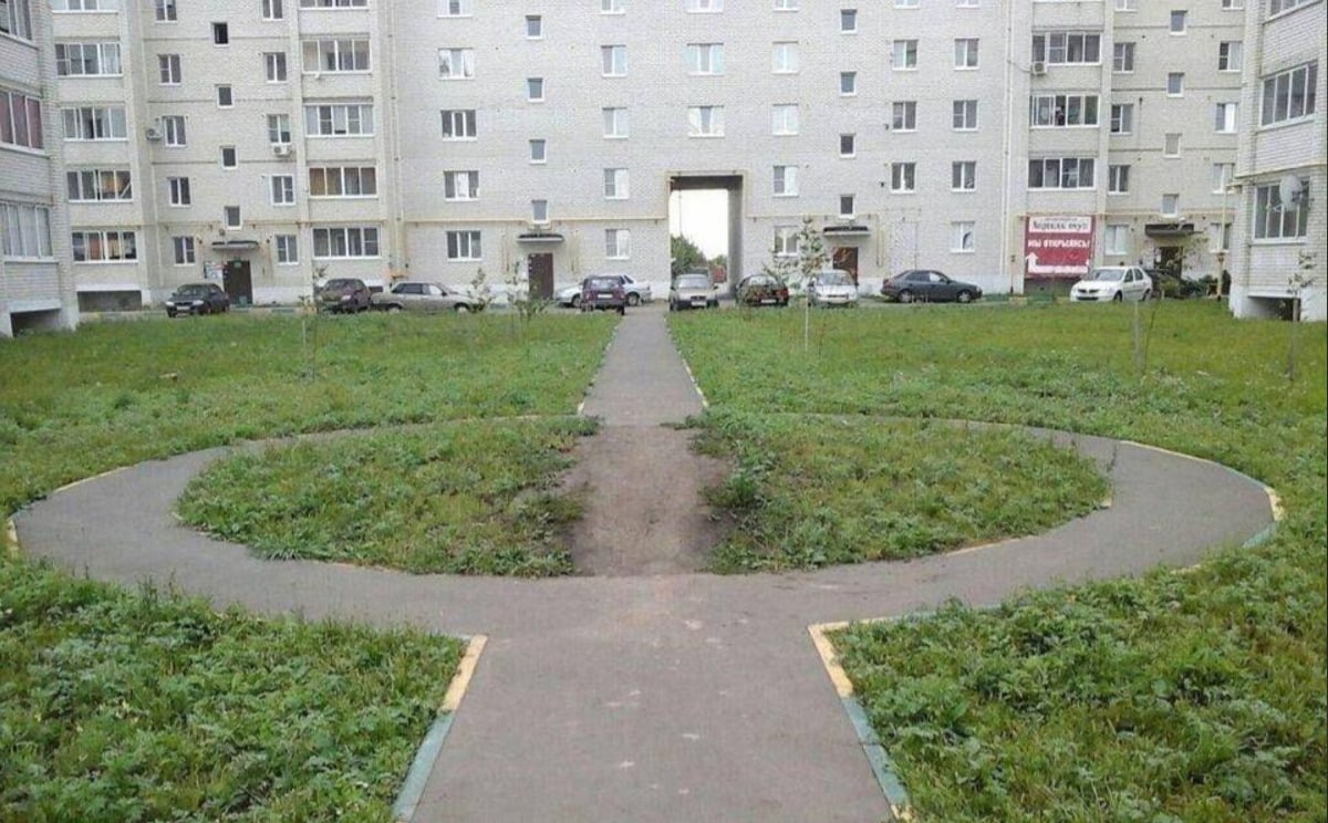 A circular pathway with a connecting sidewalk runs through a grassy courtyard surrounded by an apartment building. The path forms a circle, but a dirt shortcut cuts straight through it. Cars are parked near the building.