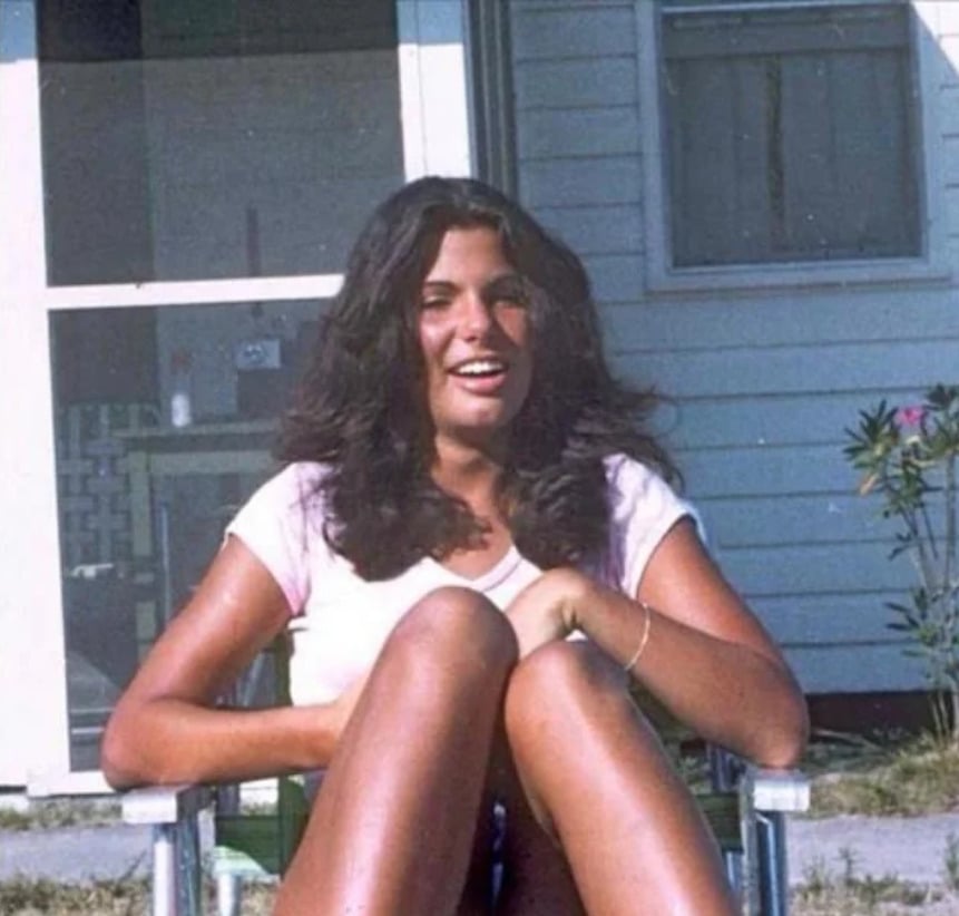 A young woman with long dark hair sits outside on a lawn chair, smiling, with her knees drawn up. She is wearing a light-colored t-shirt. A white house and some plants are visible in the background.
