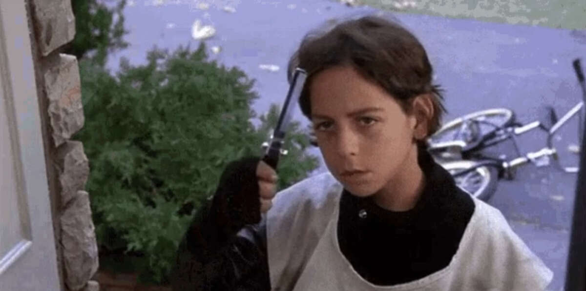 A young boy with short brown hair, wearing a white shirt and black sleeves, stands outside holding a toy gun to his head, looking serious. In the background, there is a bike lying on the ground near some greenery.