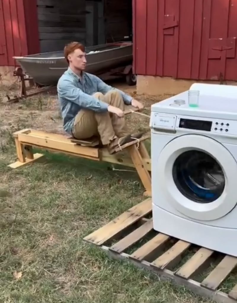A man sits on a homemade wooden rowing machine outside, using the spinning motion of a washing machine placed in front of him as resistance. Both are on a grassy area near a red barn and an old boat.