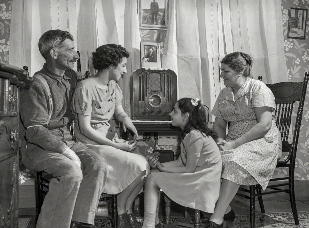 A black-and-white photo of a family of four—two adults and two girls—sitting around an old-fashioned radio, smiling and listening in a cozy, patterned living room with lace curtains.