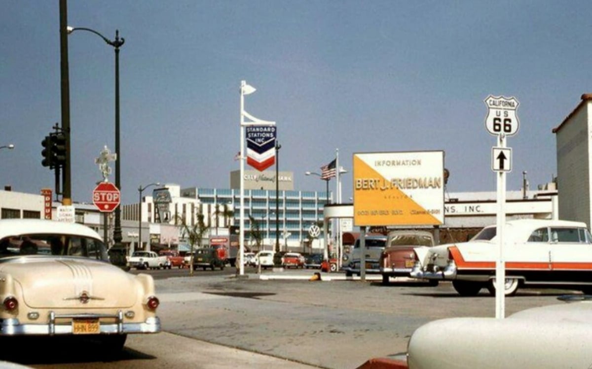 A busy 1950s street scene features vintage cars, a Chevron gas station, U.S. Route 66 signage, and a billboard for Bert L. Friedman. American flags and a large building appear in the background under a clear sky.