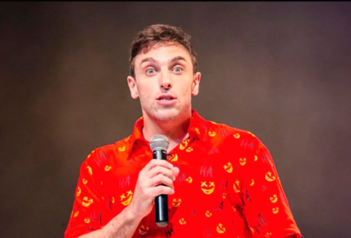 A person wearing a bright red shirt with yellow smiley faces and "HA" text holds a microphone and looks surprised or expressive, standing against a plain, neutral background.