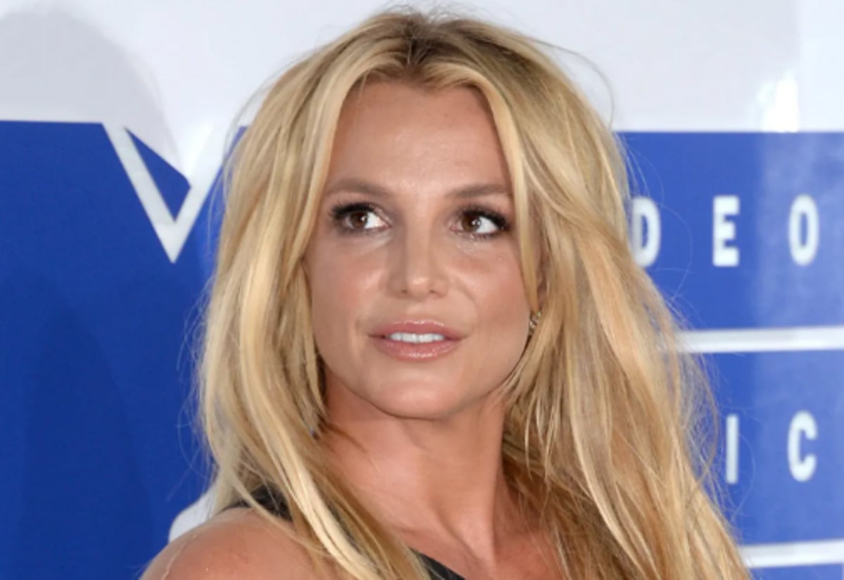 A woman with long blonde hair and a black outfit stands in front of a blue and white backdrop, looking slightly to the side with a neutral expression.