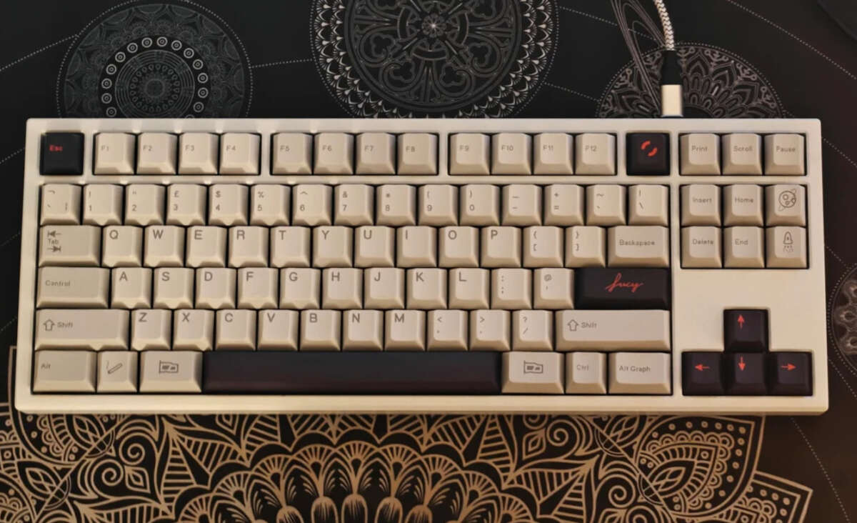A mechanical keyboard with beige keys and black legends sits on a patterned mat with intricate mandala designs in black and gold. The keyboard has some black and red accent keys and a coiled white cable attached.