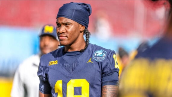 A football player in a dark blue University of Michigan uniform and head covering stands on the field, looking to the side, with blurred teammates and stadium seating in the background.
