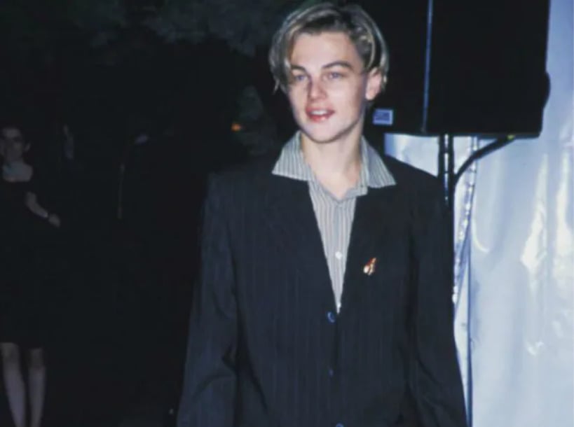 A young man with blonde hair, wearing a dark pinstripe suit and a light striped shirt, stands indoors at an event. The background is dimly lit with some indistinct people and objects.