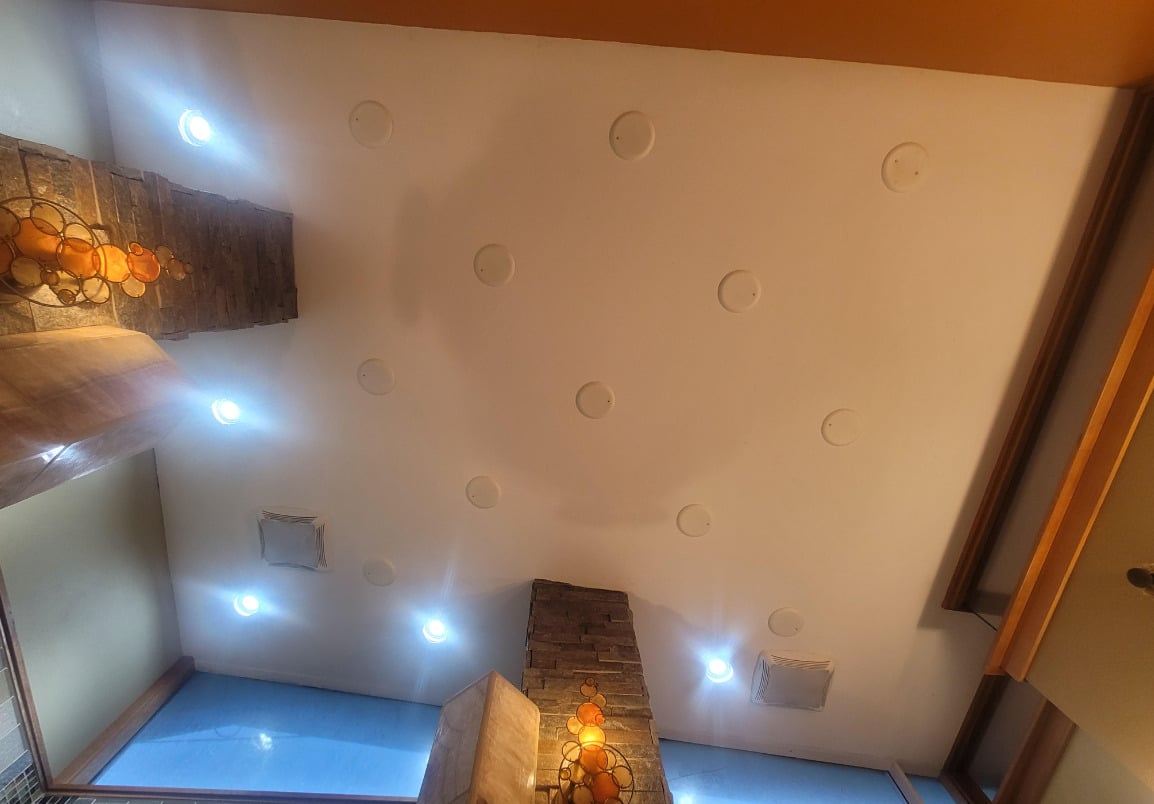 Ceiling view with recessed lights and circular accents, stone pillars, hanging amber lamps, and warm-toned architectural elements. The image is taken from a low angle, looking straight up.