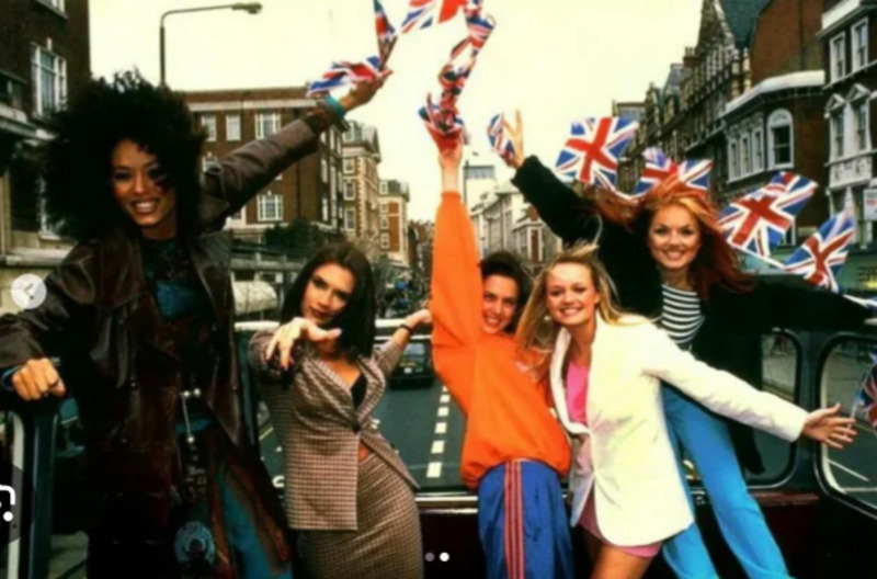 Five women pose energetically on top of a red double-decker bus, waving British flags, with city buildings in the background. They wear colorful, stylish outfits and appear joyful and lively.