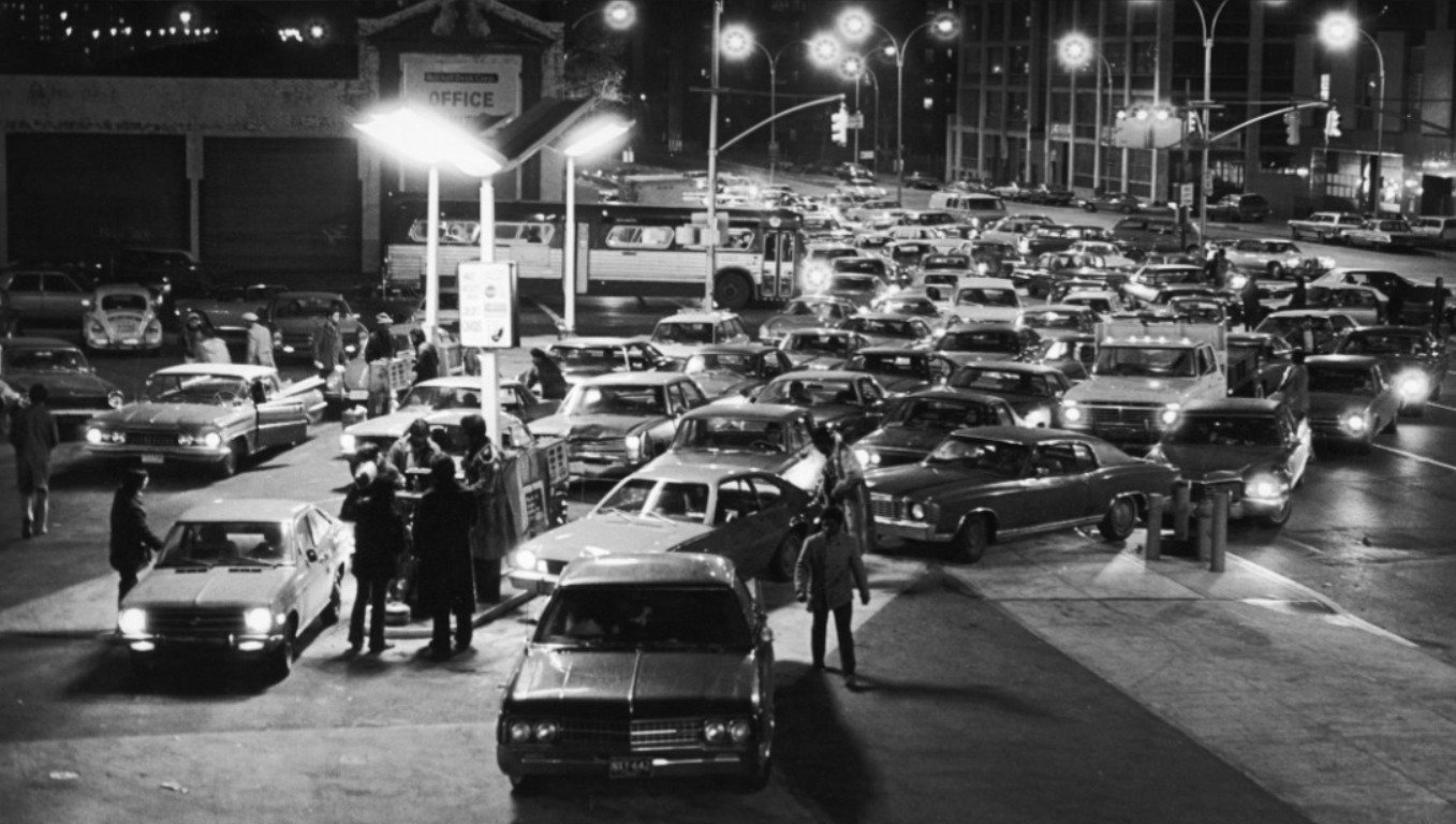 Black-and-white photo of a busy gas station at night, crowded with cars and people. Vehicles are lined up and tightly packed, with lights illuminating the scene, reflecting apparent urgency and congestion.