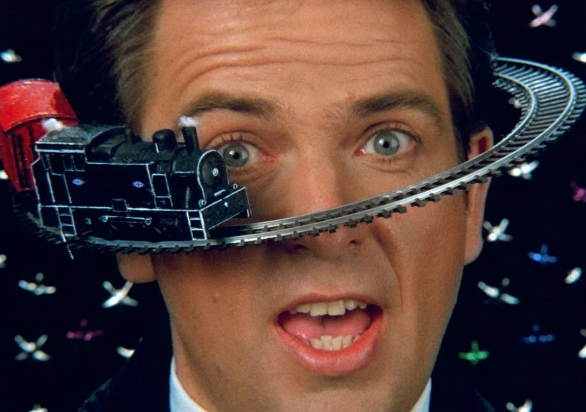 A man with blue eyes and an open mouth stands in front of a black background with colorful X shapes, as a toy train circles his head on a track that passes in front of his face and around his head.