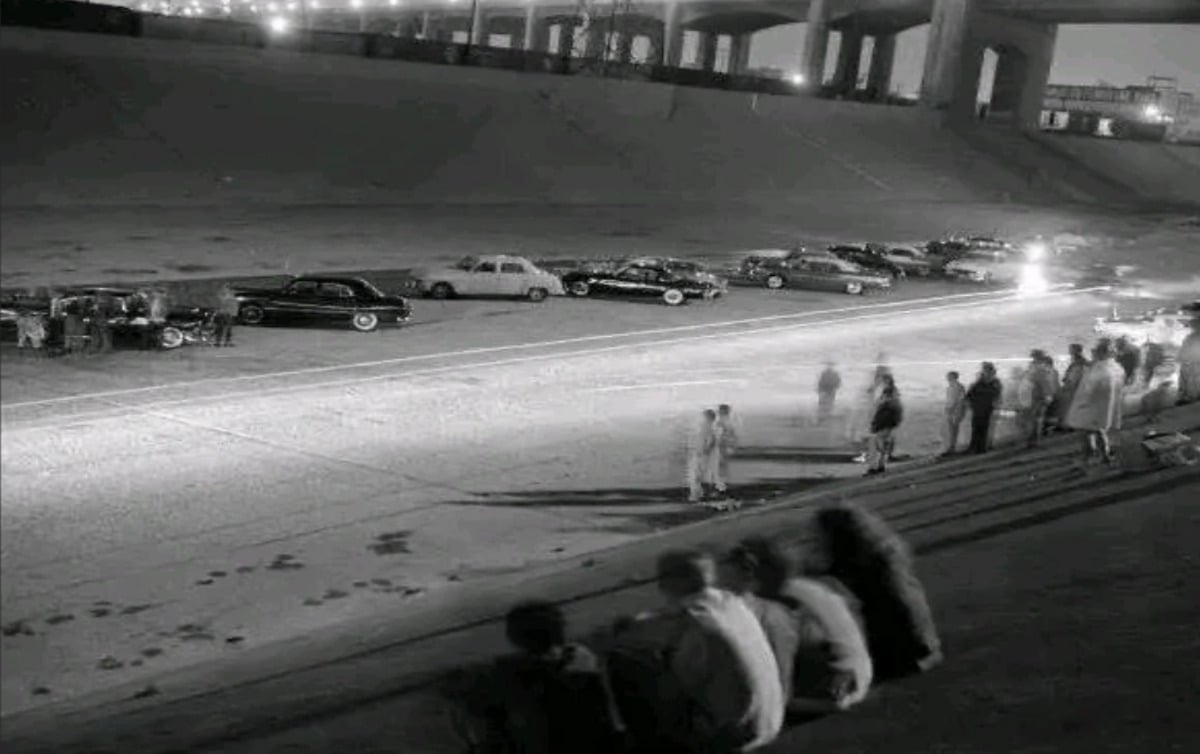 A black-and-white photo shows people gathered along the sloped sides of a wide concrete riverbed at night, watching a row of parked cars and blurred lights, possibly from a street race, under a bridge.
