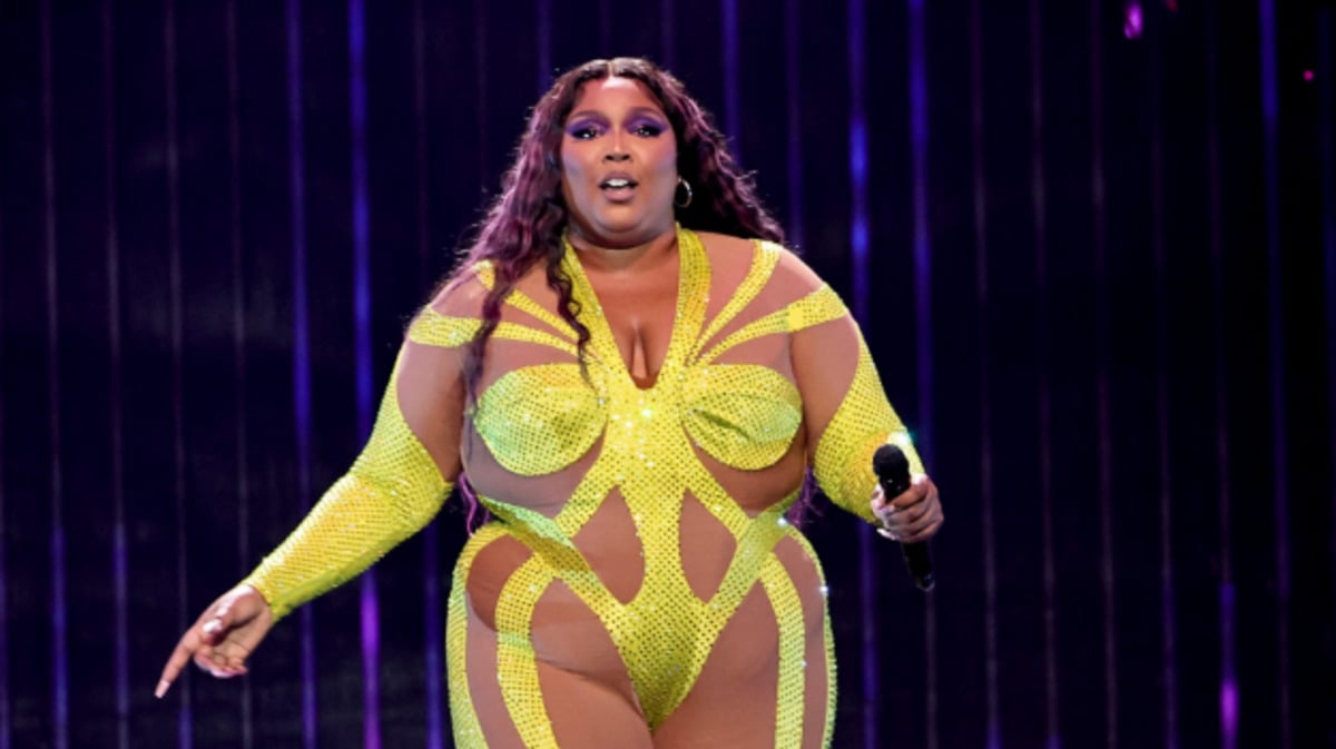 A performer in a bright yellow, sparkly bodysuit sings on stage, holding a microphone. She has long wavy hair and is standing in front of a backdrop of vertical purple lights.