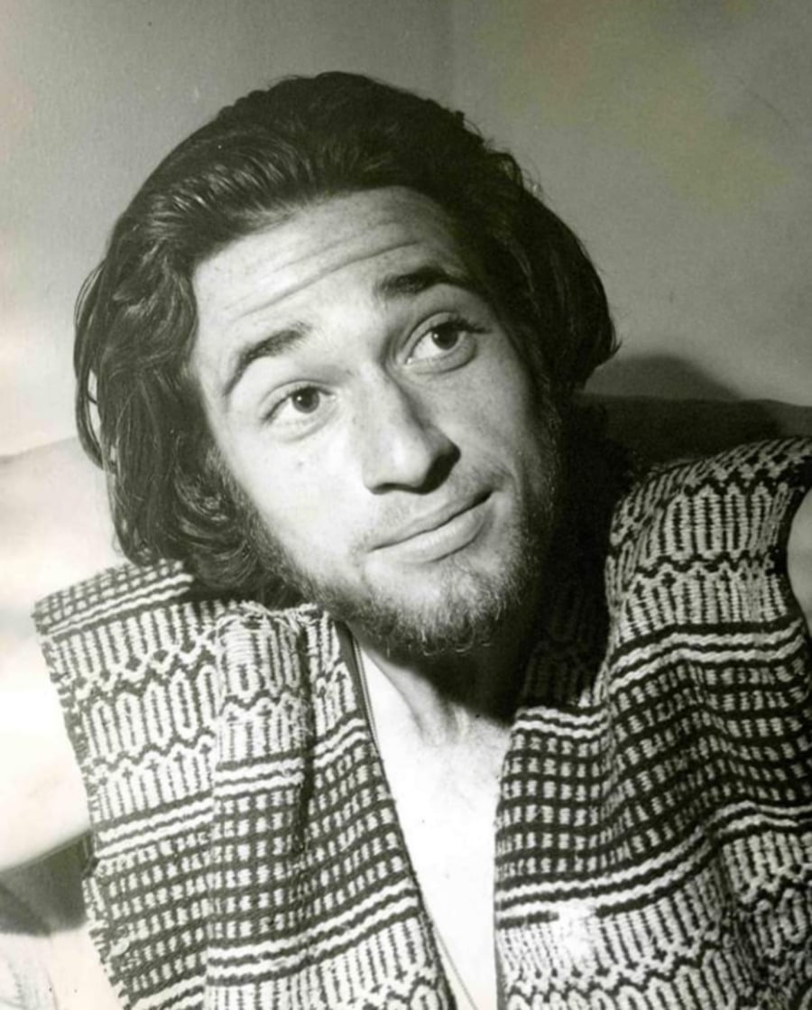 A young man with medium-length dark hair and a short beard looks slightly upward and to the side. He is wearing a textured, patterned vest and sitting in soft, indirect light. The image is in black and white.