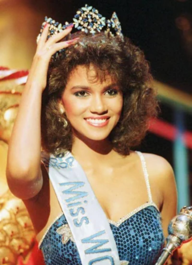 A smiling woman wearing a crown and a blue "Miss World" sash poses on stage in a sparkling blue dress, holding her hand to the crown as if adjusting it.