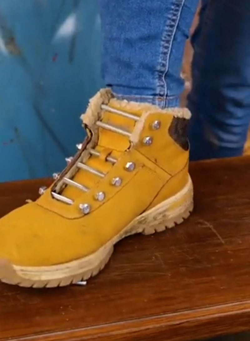 A person wearing light blue jeans and a yellow hiking boot with fur trim stands on a wooden surface. The boot’s laces are missing, and the background is blurred.