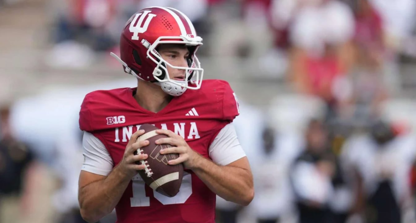 A football player in a red Indiana University uniform and helmet stands ready to throw a football during a game, looking to his left with both hands on the ball.