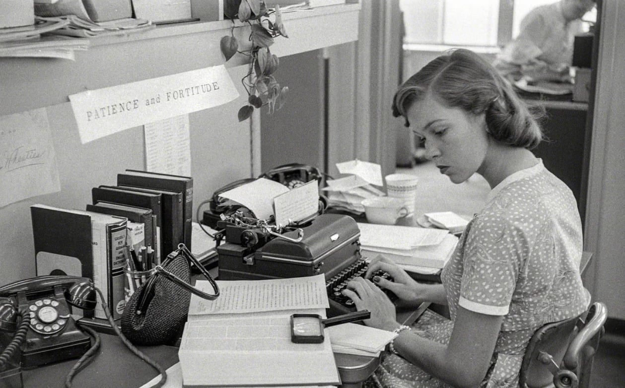 A woman in a polka-dot dress types on a typewriter at a cluttered desk with papers, books, a rotary phone, and a coffee cup. A sign on the wall reads “Patience and Fortitude.” Another person sits in the background.