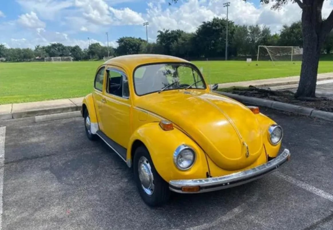 A bright yellow vintage Volkswagen Beetle is parked in a lot next to a grassy field with soccer goals, under a partly cloudy sky.