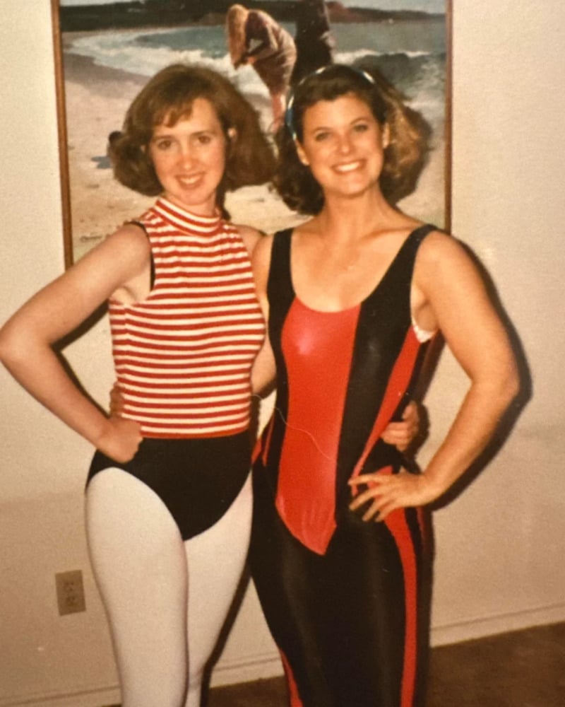 Two women wearing colorful 1980s-style leotards stand smiling with their arms around each other indoors, in front of a painting of a beach scene.