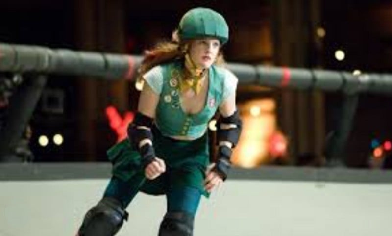 A woman in green roller derby gear, including a helmet, knee and elbow pads, skates on an indoor track with a focused expression. She wears a sleeveless top with several pins and patches.
