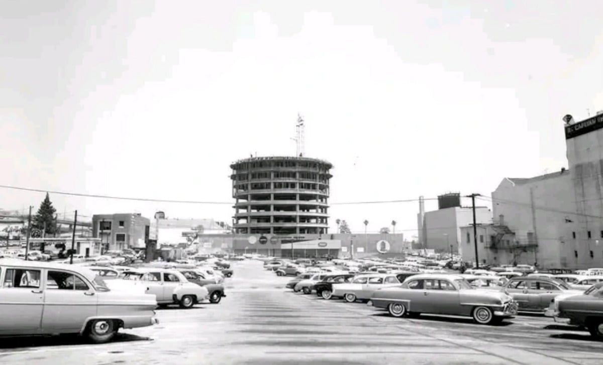 Black-and-white photo of a large parking lot filled with vintage cars. In the background, a round building is under construction, surrounded by other low-rise buildings, power lines, and signs.