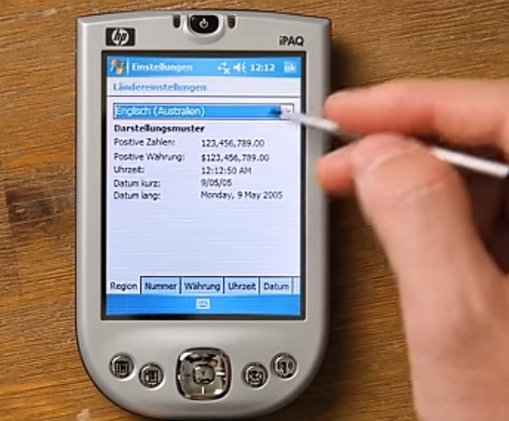 A hand uses a stylus to interact with a silver HP iPAQ handheld device displaying settings in German and English on its screen. The device rests on a wooden surface.