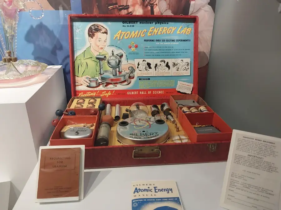 A vintage “Atomic Energy Lab” science kit is displayed in an open case, containing instruments, sample jars, manuals, and instructional booklets. The colorful box lid features a child using lab equipment and scientific illustrations.