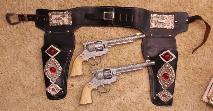 A black leather cowboy gun belt with silver buckles and red gem accents, holding two ornate silver toy revolvers with ivory-colored handles, laid out on a beige carpet.