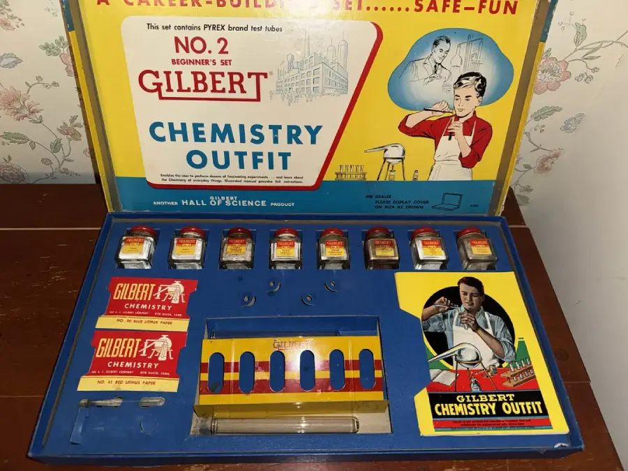 A vintage Gilbert Chemistry Outfit set displayed open, showing various labeled chemical containers, test tubes, instruction booklets, and tools, with colorful graphics and illustrations on the inside lid and box.
