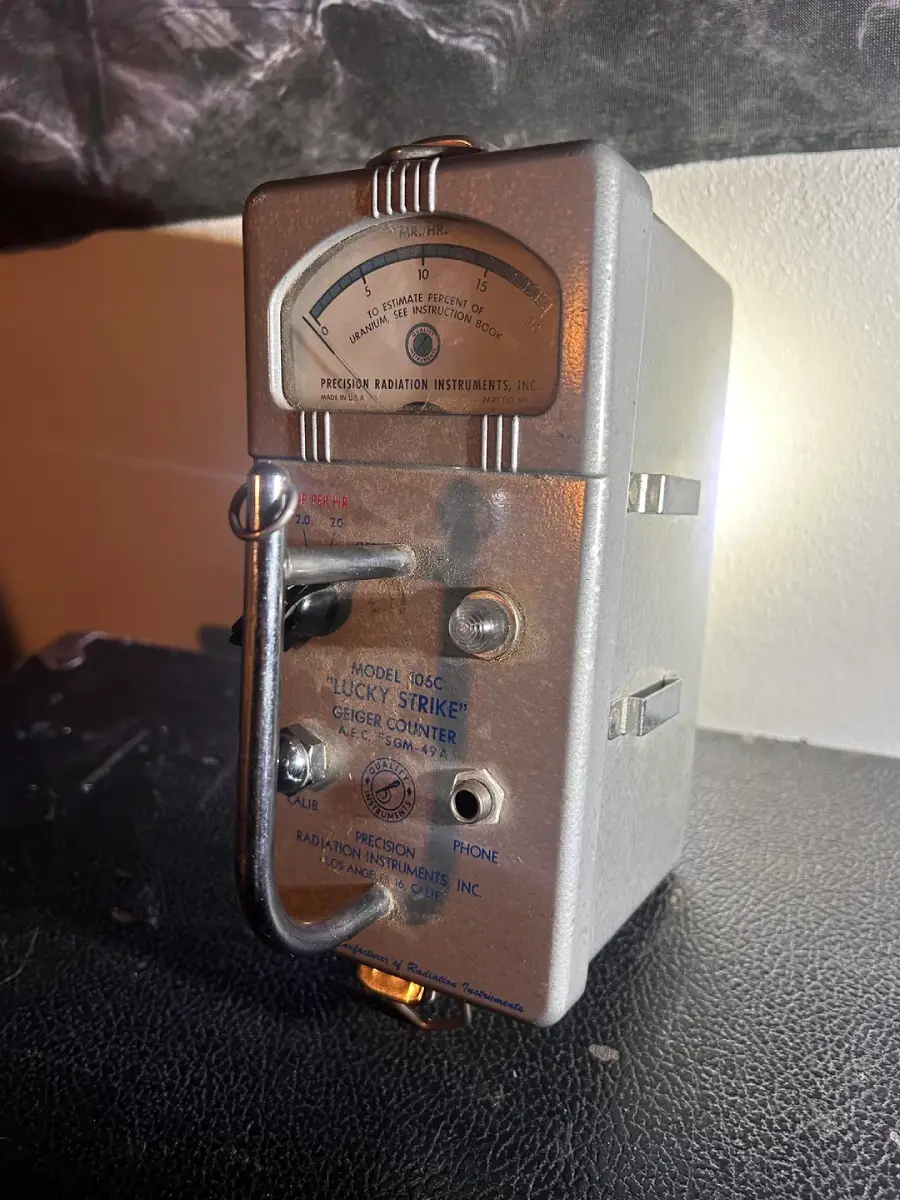A vintage silver Geiger counter labeled "Lucky Strike, Model 106C" sits on a dark surface. It features a needle gauge, switches, and dials on the front panel, with text indicating radiation measurement.