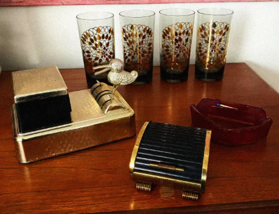 A set of four patterned glasses, a red ashtray, a gold and black cigarette case, and a gold retro lighter sit on a wooden table.