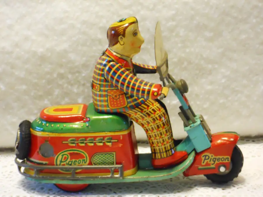 A colorful vintage tin toy of a man riding a red, green, and blue scooter labeled "Pigeon." The man wears a plaid suit and sits upright, holding the handlebars. The background is a neutral textured surface.
