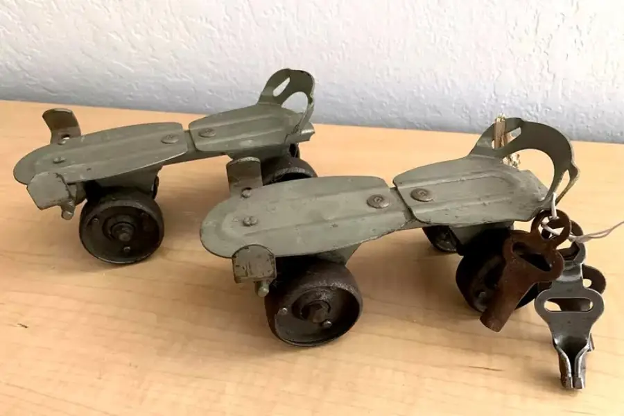 A pair of vintage metal roller skates with adjustable footplates and large front wheels sits on a wooden surface. A set of keys is attached to one skate for adjusting the size.