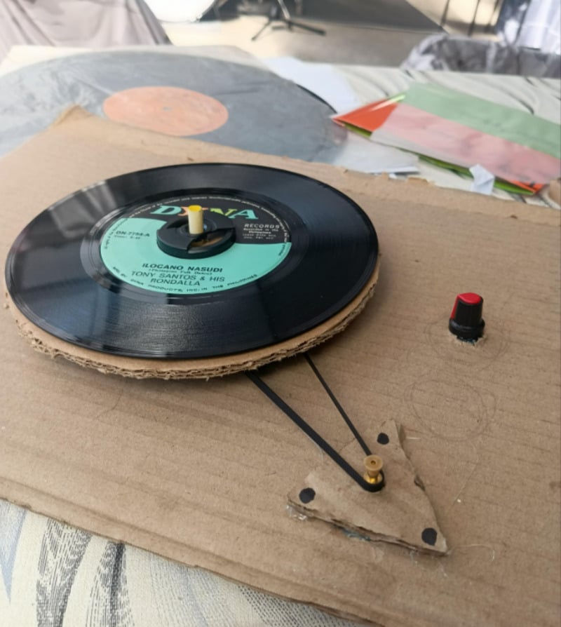 A homemade record player made from cardboard, playing a vinyl record with visible rubber bands, a small black knob, and a red button on a cardboard base. Papers and another record are in the background.