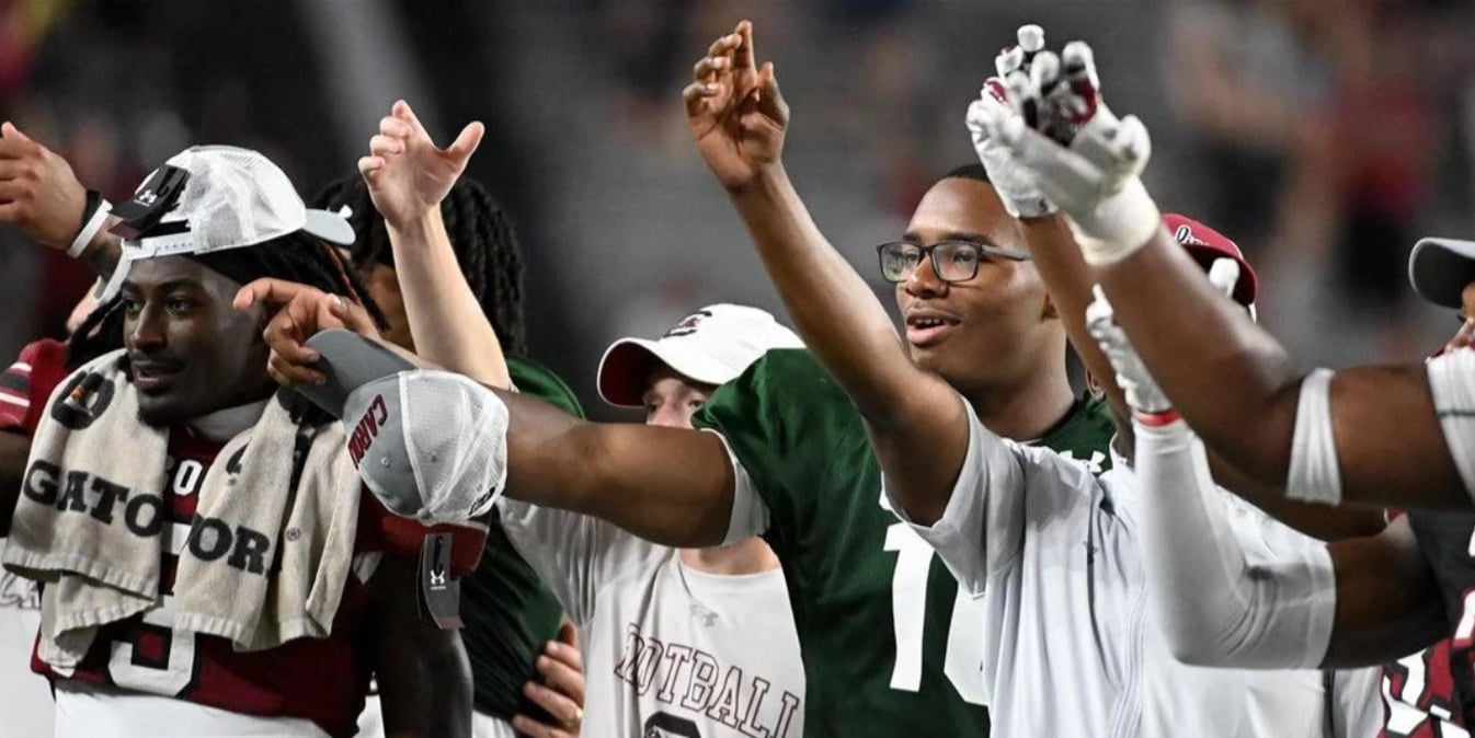 A group of football players, some in uniform and some in warm-ups, stand arm in arm, smiling and raising their hands in celebration on the field after a game.