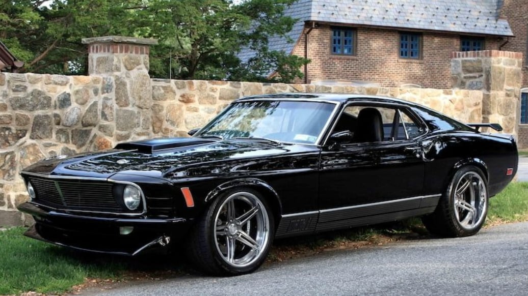 A shiny black classic muscle car, likely a late 1960s or early 1970s Ford Mustang, is parked on a street beside a stone wall and trees, with a brick building in the background.