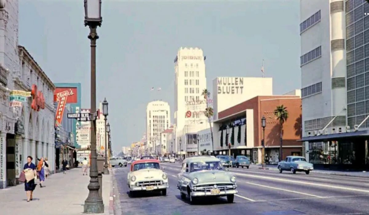A street scene from the 1950s shows vintage cars driving down a wide avenue lined with palm trees, pedestrians on sidewalks, and tall buildings with store signs in a sunny, urban setting.