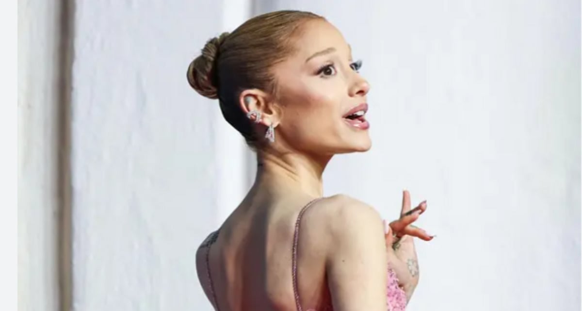 A woman with her hair in a bun, wearing a sparkly pink dress and earrings, poses with her head turned to the side against a light background.