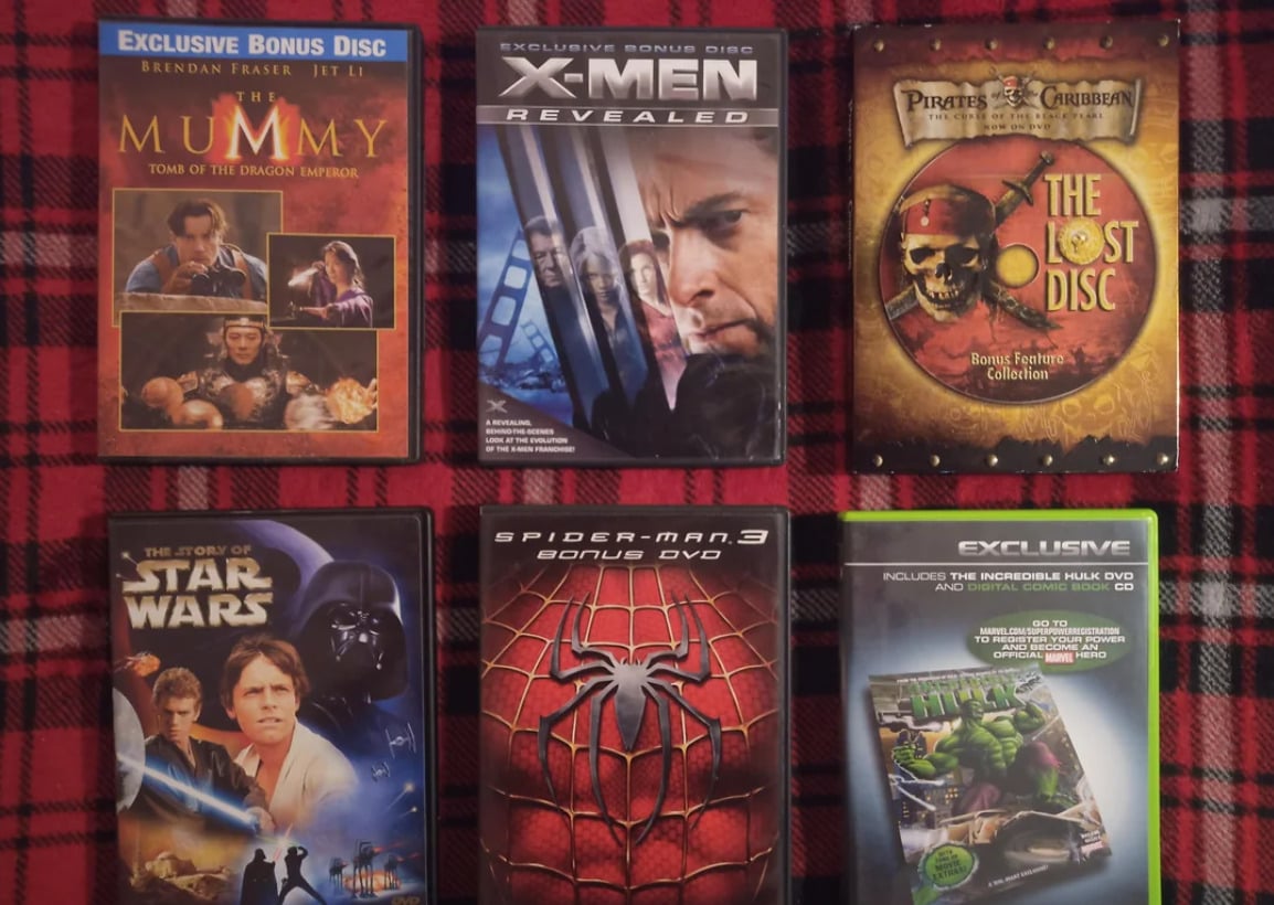 Six DVD and CD bonus discs are arranged on a red plaid surface. Titles include The Mummy, X-Men Revealed, Pirates of the Caribbean, Star Wars, Spider-Man 3, and The Incredible Hulk.