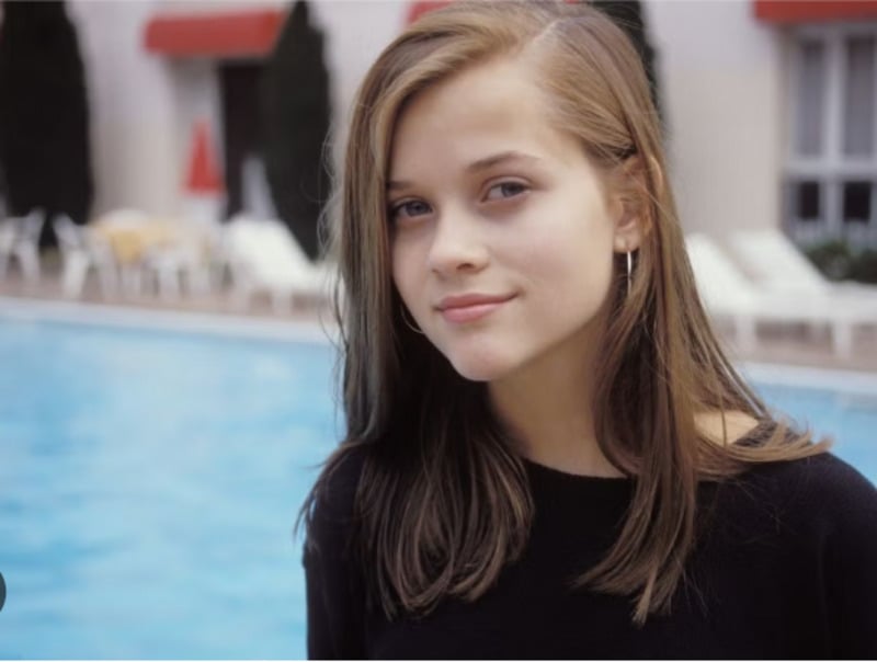 A young woman with straight, light brown hair and hoop earrings smiles subtly while standing by an outdoor swimming pool. White lounge chairs and red awnings are visible in the background.