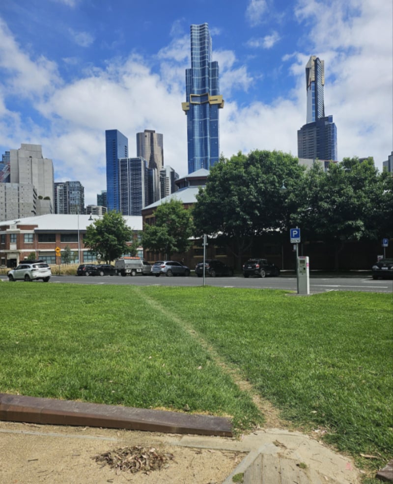 A grassy park area with a dirt path leads toward a street with parked cars, trees, and several modern skyscrapers in the background under a partly cloudy sky.