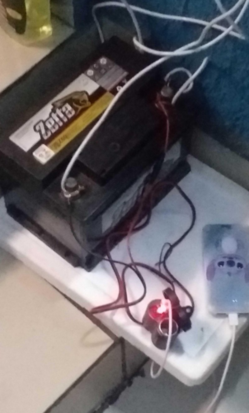 A black car battery labeled "Zebra" is connected with red and black wires to a small device with a glowing red light and a plugged-in mobile phone charger on a white surface.