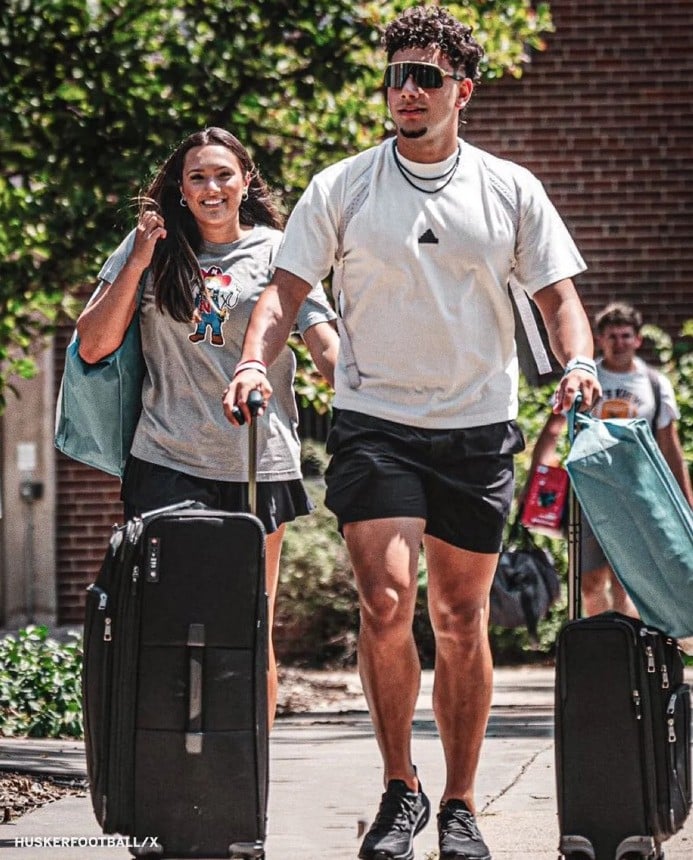 Two young adults, both wearing light-colored shirts and black shorts, walk outdoors on a sunny day while pulling large suitcases. They are smiling, and trees and a brick building are visible in the background.