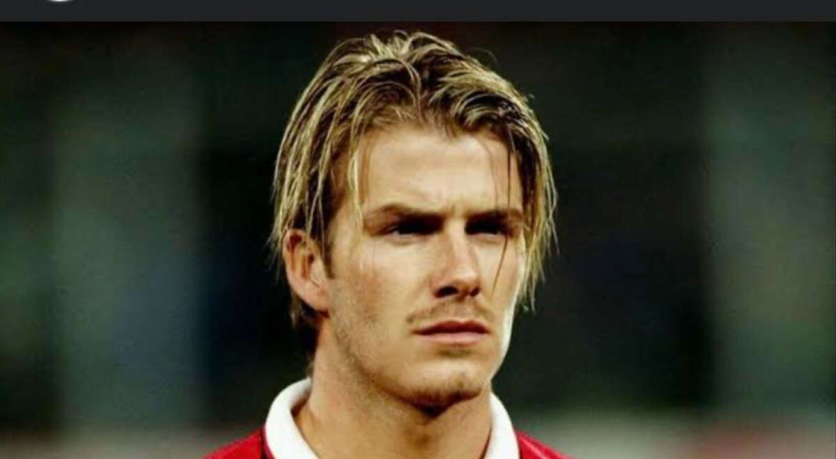 A young man with blonde, tousled hair wearing a red sports jersey with a white collar, looking serious and focused against a blurred background.