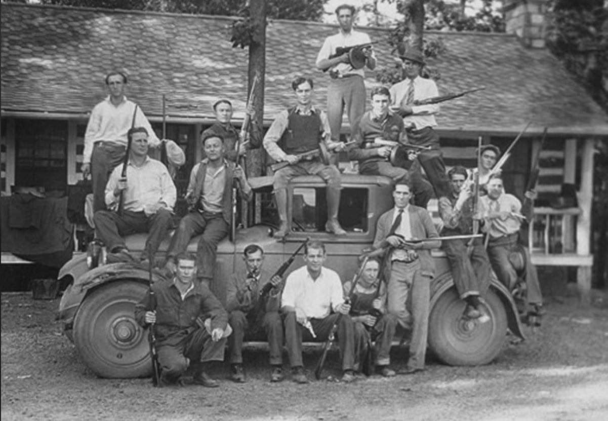 A group of men posing with rifles and shotguns on and around an old car in front of a rustic building, likely from the early 20th century. Some men are standing, others are sitting, and many are holding firearms.