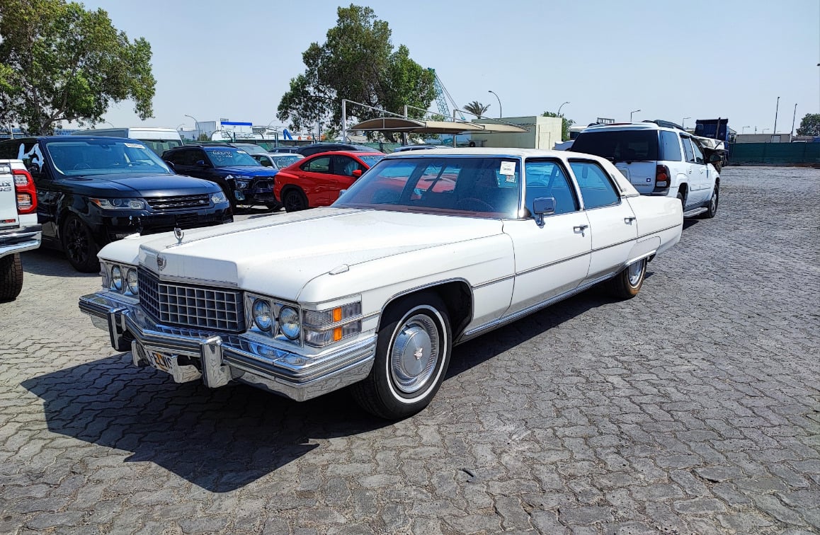 A vintage white Cadillac sedan is parked on a cobblestone lot among modern black and white SUVs and cars, with trees and buildings in the background under a clear sky.
