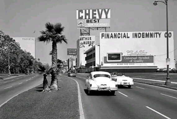 A black-and-white photo of a mid-20th-century boulevard with vintage cars, palm trees, and large advertising billboards for Chevy, Financial Indemnity Co., Arthur Murray, and Cadillac.