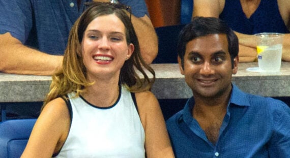 A woman and a man sit close together, smiling at an event. The woman has long brown hair and wears a sleeveless white top; the man has short dark hair and wears a blue button-up shirt. People and drinks are in the background.
