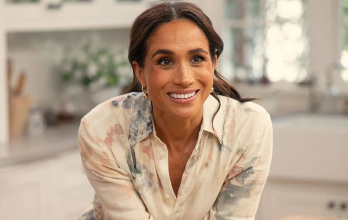 A woman with long dark hair, wearing a light-colored, floral blouse, smiles warmly in a bright kitchen with white cabinets and a blurred background.